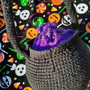Op de afbeelding: Een zwarte gehaakte tas met een paars gehaakt item erin, tegen een zwarte stof met patronen van Halloween-lolly's, snoepma&iuml;s en pompoengezichten. De tas heeft een riem en is ontworpen om items te dragen.