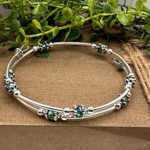 May include: A silver-toned bangle bracelet with two parallel bands. The bracelet is adorned with iridescent green and silver beads. The beads are interspersed with smaller silver accents. The bracelet is displayed on a wooden surface with greenery in the background.