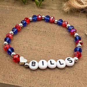 May include: A red, white, and blue beaded bracelet with a silver heart charm and the word "BILLS" spelled out in white beads.