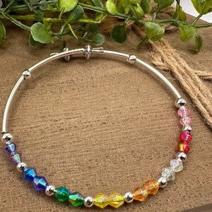 May include: A silver bangle bracelet with a rainbow of faceted beads. The beads transition from blue to red, with silver accent beads. The bracelet is displayed on a wooden surface.