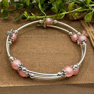 May include: A silver bangle bracelet with two rows of pink beads. The beads have a sparkly finish and are accented with silver charms.