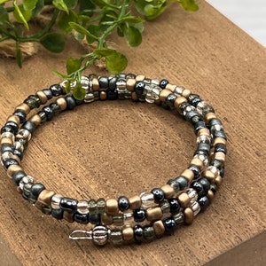 May include: A multi-strand beaded bracelet with gold, silver, and black beads. The bracelet is wrapped around a wooden surface.