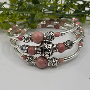 May include: A multi-strand wrap bracelet featuring smooth pink stone beads, silver-toned metal spacers with intricate floral and swirl patterns, and small faceted grey beads. The design consists of three coiled loops with a polished metallic finish.