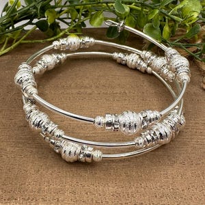 May include: A set of three silver-toned, spiral bracelets. Each bracelet features textured, beaded accents. The bracelets are displayed on a brown surface with green foliage in the background. The bracelets are a stylish accessory.