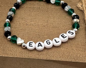 May include: A green and black beaded bracelet with a silver heart charm and white letter beads spelling "EAGLES".