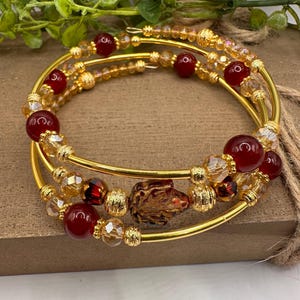 May include: A multi-strand gold-tone wire wrap bracelet featuring deep red spherical beads, translucent amber-colored faceted crystals, and small textured gold-tone spacer beads. A central dark red leaf-shaped bead serves as a focal point.