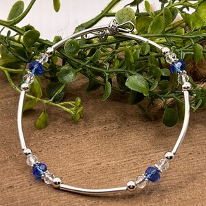 May include: A silver bangle bracelet with a clasp closure. The bracelet is adorned with clear and blue crystal beads.