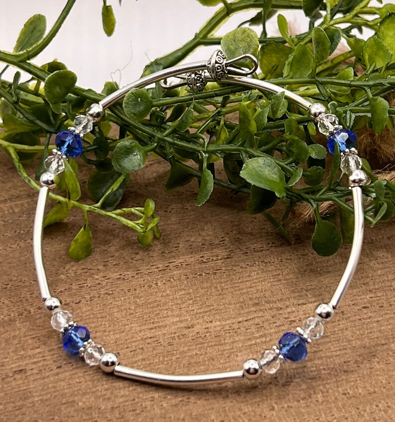May include: A silver bangle bracelet with a clasp closure. The bracelet is adorned with clear and blue crystal beads.