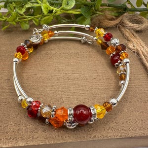 May include: A multi-strand silver-tone wire bangle bracelet featuring an assortment of faceted crystal beads in shades of red, orange, yellow, and brown, accented with small silver-tone metal spacers and a single large smooth dark red bead.