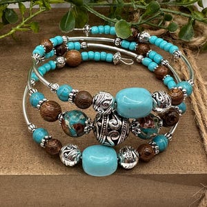 May include: A multi-strand wrap bracelet featuring turquoise-colored beads, dark brown wood-grain beads, and silver-toned metal spacers with intricate scrollwork patterns. The design incorporates smooth, polished turquoise stones and textured beads.