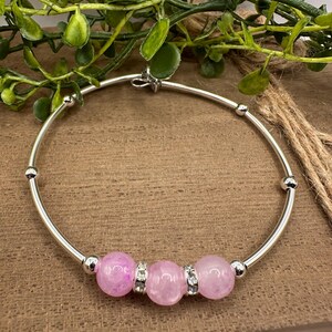 May include: A silver-toned bangle bracelet featuring three round, translucent pink beads separated by two small silver-toned spacers encrusted with clear crystals. The bracelet includes additional small silver-toned metal beads along the circular band.