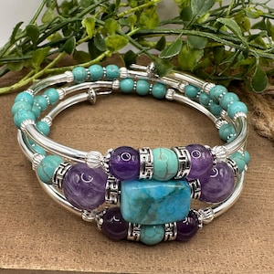 May include: A silver-toned, multi-strand bracelet with turquoise and amethyst beads. The bracelet features a large, rectangular turquoise stone at the center, surrounded by smaller beads and silver accents. The design is set against a wooden surface.