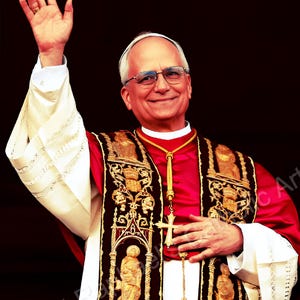 Può includere: Un uomo con una veste religiosa rossa e nera con ricami dorati, una croce bianca e occhiali, che saluta con la mano destra. L'immagine ha uno sfondo scuro.