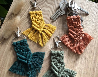 May include: Four handmade macrame keychains with fringe in yellow, green, orange, and sage green. Each keychain has a silver metal ring.