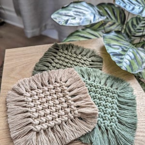 May include: Three handmade macrame coasters in beige and green with fringe detailing. The coasters are made with cotton cord and are perfect for adding a touch of bohemian style to your home.