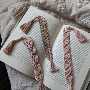 Pode incluir: Três marcadores de livro de macramé rosa e castanho com contas de madeira e borlas. Os marcadores de livro são feitos à mão e apresentam um padrão de nó único.