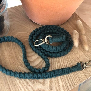 Puede incluir: Una correa de cuerda de algodón verde oscuro con un clip de metal plateado. La correa está enrollada sobre una superficie de madera.