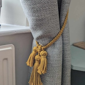 May include: A mustard yellow tassel tieback for curtains. The tieback is made of a thick, braided rope with three tassels at the end. The tieback is tied around a grey curtain.