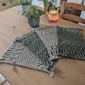 Könnte beinhalten: Drei gehäkelte Tischsets in einem beige-grünen Farbschema. Die Tischsets sind übereinander gelegt, wobei das beige Tischset unten und die grünen Tischsets oben liegen. Die Tischsets sind mit einer Makramee-Technik hergestellt und haben einen Fransenrand.