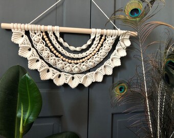 May include: A white macrame wall hanging with wooden beads, featuring a crescent shape with multiple layers of intricate knotting. The hanging is displayed against a dark gray background with peacock feathers.