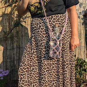 May include: A person wearing a black t-shirt with a graphic print and a leopard print skirt. A pink crocheted bottle holder with white beads is attached to the skirt and holds a clear glass bottle.