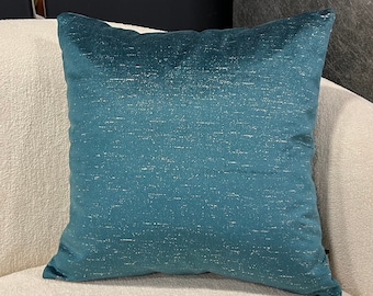 Puede incluir: Un cojín de sofá azul turquesa con un brillo sutil, apoyado en un sofá blanco.
