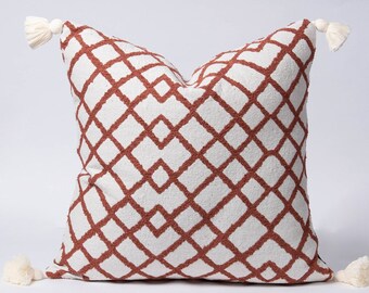 May include: Square decorative pillow with a white background and a geometric diamond pattern in a rust color. The pillow has four corners, each adorned with a small, cream-colored tassel. The pillow is designed for home decor.