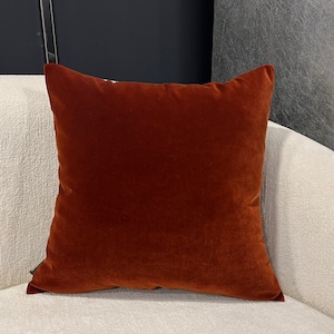 Op de afbeelding: Een vierkant kussen van fluweel in roestkleur. Het kussen ligt op een wit oppervlak.