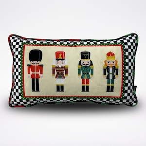 Puede incluir: Una almohada decorativa con un borde a cuadros blanco y negro y un fondo crema. La almohada presenta cuatro soldados cascanueces en diferentes poses y colores.