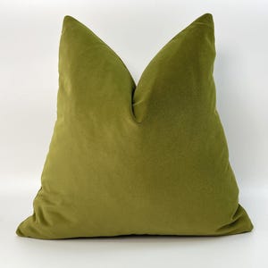 May include: A square throw pillow featuring a solid olive green velvet cover. The pillow has a soft, plush texture with a slight sheen and is shaped with a distinct peak at the top center, creating a structured appearance against a plain background.