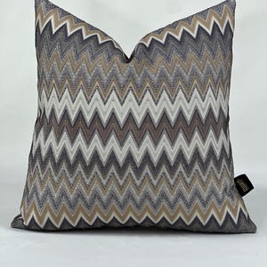 Op de afbeelding: Een vierkant sierkussen met een herhalend chevronpatroon in tinten donkergrijs, lichtgrijs, beige en wit. De stof heeft een textuur en aan de rechterkant zit een klein zwart stoffen label met de tekst Mood Home bevestigd.