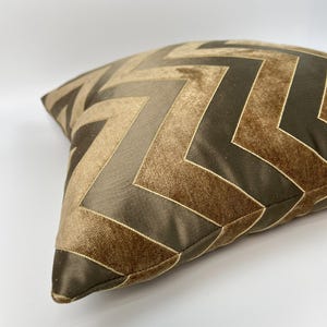 May include: A square throw pillow featuring a repeating chevron pattern in alternating shades of dark brown and light tan. The design consists of alternating panels of smooth, satin-like fabric and a soft, textured velvet material with thin piping.