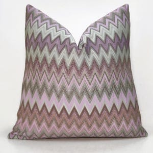 Może przedstawiać: Dekoracyjna poduszka z wzorem w szewrony w odcieniach różu, szarości i brązu. Zygzakowaty wzór tworzy teksturowany wygląd. Poduszka ma kwadratowy kształt i wydaje się być wykonana z miękkiej tkaniny.