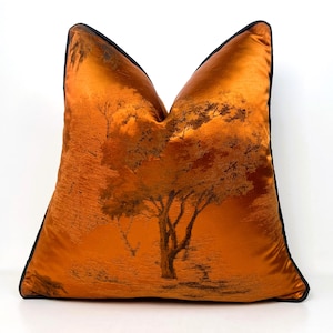Può includere: Un cuscino decorativo arancione con bordatura nera. Il cuscino presenta un design a paesaggio di alberi nelle tonalità dell'arancione e del marrone. Il cuscino è di forma quadrata e sembra essere realizzato in un materiale simile al raso.
