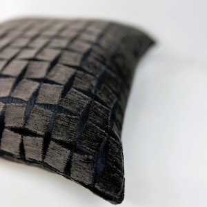Puede incluir: Un cojín cuadrado decorativo con un patrón texturizado y tejido. El cojín presenta una combinación de colores gris oscuro y negro, con un diseño geométrico repetitivo. El cojín está hecho de un material suave y afelpado, y probablemente se utiliza para la decoración del hogar.