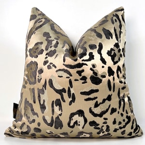 May include: A square decorative pillow with a gold background and a black and gray leopard print pattern. The pillow has a soft, textured appearance and is likely made of a luxurious fabric. The pillow is a home decor item.