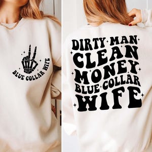 May include: A cream colored sweatshirt with a black skeleton hand peace sign and the text "Blue Collar Wife" on the front. The back of the sweatshirt has the text "Dirty Man Clean Money Blue Collar Wife" in black.