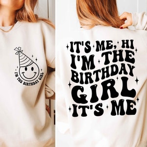 May include: A cream-coloured sweatshirt with a black smiley face wearing a party hat and the text "I'm the birthday girl" on the front. The back of the sweatshirt has the text "It's me, hi I'm the birthday girl it's me" in black.