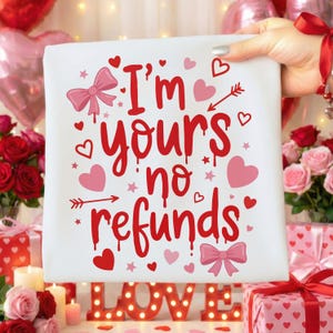 May include: A white square fabric with the text "I'm yours no refunds" in red, surrounded by pink hearts, bows, and stars. The fabric is held up by a hand. The theme is Valentine's Day.