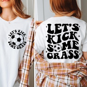 K&ouml;nnte beinhalten: Wei&szlig;es T-Shirt mit dem Text "Let's Kick Some Grass" in schwarzer Retro-Schrift mit einem Fu&szlig;ball-Motiv. Die R&uuml;ckseite des Shirts hat den gleichen Text und das gleiche Motiv in gr&ouml;&szlig;erer Gr&ouml;&szlig;e.