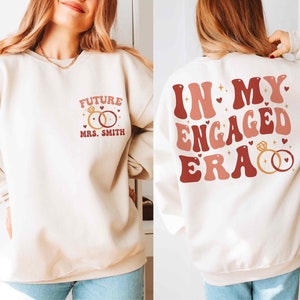 May include: A white sweatshirt with a red and pink graphic on the back that says "In My Engaged Era" with two gold rings. The front of the sweatshirt has a red and pink graphic that says "Future Mrs. Smith" with two gold rings.