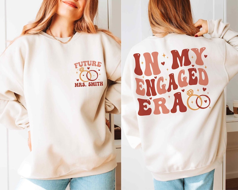 May include: A white sweatshirt with a red and pink graphic on the back that says "In My Engaged Era" with two gold rings. The front of the sweatshirt has a red and pink graphic that says "Future Mrs. Smith" with two gold rings.