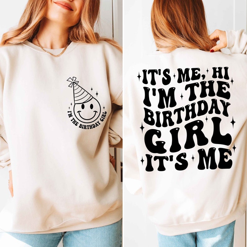 May include: A cream-coloured sweatshirt with a black smiley face wearing a party hat and the text "I'm the birthday girl" on the front. The back of the sweatshirt has the text "It's me, hi I'm the birthday girl it's me" in black.