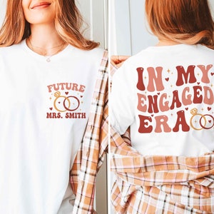 May include: Two white t-shirts with a pink and red graphic design. The first shirt says "Future Mrs. Smith" with a ring design. The second shirt says "In My Engaged Era" with a ring design.