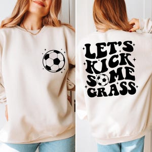 K&ouml;nnte beinhalten: Ein cremefarbener Pullover mit einem schwarzen Fu&szlig;ball-Motiv auf der Vorderseite und dem Text "Let's Kick Some Grass" auf der R&uuml;ckseite.