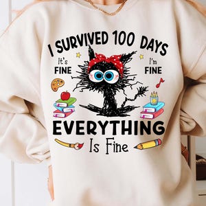 May include: A white sweatshirt with a black cat illustration and the text "I survived 100 days Everything is fine". The cat is wearing a red and white polka dot headband. The text is surrounded by school supplies like paintbrushes, pencils, and books.