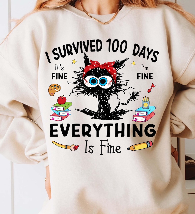 May include: A white sweatshirt with a black cat illustration and the text "I survived 100 days Everything is fine". The cat is wearing a red and white polka dot headband. The text is surrounded by school supplies like paintbrushes, pencils, and books.