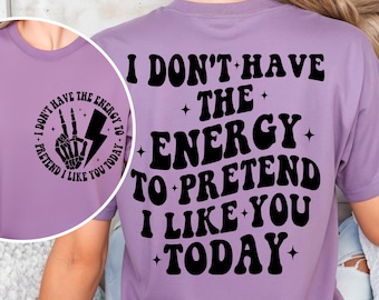 May include: A lilac-colored t-shirt with the text "I DON'T HAVE THE ENERGY TO PRETEND I LIKE YOU TODAY" in black. A circular graphic on the front features a skeleton hand, lightning bolt, and the text "I DON'T HAVE THE ENERGY TO PRETEND I LIKE YOU TODAY."