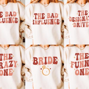 Puede incluir: Una colección de seis sudaderas de cuello redondo color crema con varias frases en una fuente ondulada de tonos rojos y rosas. El texto incluye "THE BAD INFLUENCE", "THE DESIGNATED DRIVER", "THE CRAZY ONE", "BRIDE" con un icono de anillo y "THE FUNNY ONE".