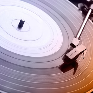 Op de afbeelding: Close-up van een vintage platenspeler met een vinylplaat. De plaat is grijs met concentrische groeven en de toonarm is boven de plaat geplaatst. De afbeelding heeft een warme, oranje en paarse gradi&euml;nt.